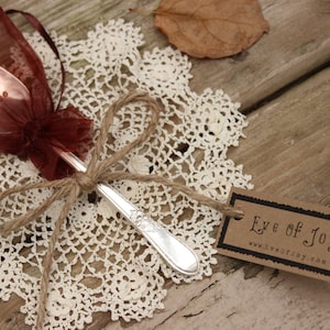 May include: A silver spoon with an inscription on the bowl, tied with twine and placed on a white crocheted doily. The spoon is wrapped in a burgundy sheer fabric and a brown paper tag with the text "Eye of Joy" and a website address.