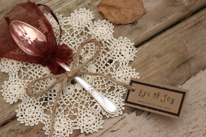 May include: A silver spoon with an inscription on the bowl, tied with twine and placed on a white crocheted doily. The spoon is wrapped in a burgundy sheer fabric and a brown paper tag with the text "Eye of Joy" and a website address.