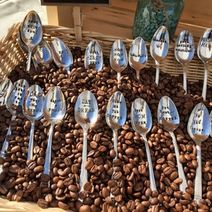 May include: A collection of silver spoons with various coffee-themed sayings engraved on them, displayed in a wicker basket filled with coffee beans.