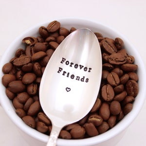 May include: A silver spoon with the words "Forever Friends" engraved on it, surrounded by coffee beans in a white bowl.