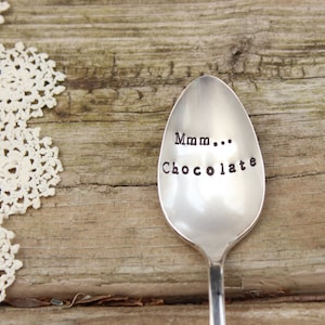 May include: A silver spoon with the words "Mmm... Chocolate" stamped on the bowl. The spoon is set against a rustic wooden background, with a piece of white lace in the upper left corner.