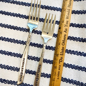 May include: Two silver forks with the words "Dinglehopper" engraved on the handles. The forks are on a white and blue striped surface with a wooden ruler that says "A good rule "Do Unto Others As You Would Have Them" in the background.