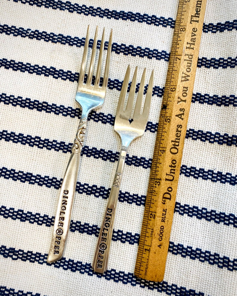 May include: Two silver forks with the words "Dinglehopper" engraved on the handles. The forks are on a white and blue striped surface with a wooden ruler that says "A good rule "Do Unto Others As You Would Have Them" in the background.