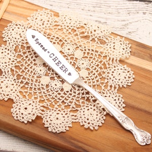 Può includere: Coltello da burro d'argento con le parole "Spread Cheer" incise sul manico. Il coltello è appoggiato su un pizzo bianco all'uncinetto.