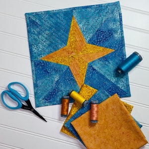 May include: A blue and yellow fabric quilt block with a star design. The quilt block is surrounded by spools of thread and a pair of scissors.