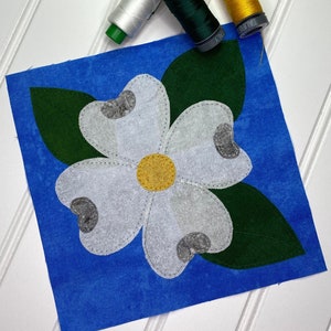 May include: A blue fabric square with a white flower with a yellow center and green leaves. The flower has five petals and is surrounded by three green leaves. There are three spools of thread in the background: white, green, and yellow.
