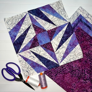 May include: A purple and white quilt block with a geometric pattern. The block is made of fabric scraps and is ready to be sewn into a larger quilt. There are also two spools of thread and a pair of scissors on the white surface.