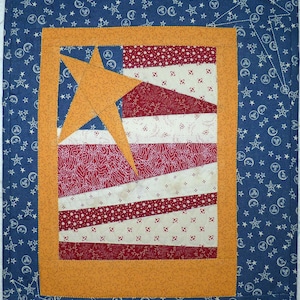 Pode incluir: Um bloco de colcha patriótico emoldurado com um design de estrela vermelho, branco e azul. O bloco de colcha é cercado por um tecido azul com estrelas brancas.