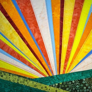 Könnte beinhalten: Ein farbenfrohes, abstraktes Quilt mit einem Sonnenaufgangsdesign. Das Quilt zeigt strahlenförmige Streifen in Orange, Gelb, Rot und Blau, mit einer grünen Landschaft am unteren Rand. Der Stoff ist mit sichtbaren Nähten zusammengenäht.