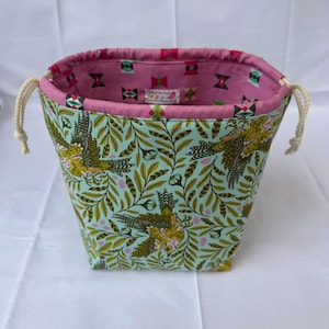 May include: A fabric drawstring bag with a light blue exterior featuring a bird and leaf pattern in shades of green and gold. The interior is pink with a geometric design. The bag has a drawstring closure with cream-colored cords.