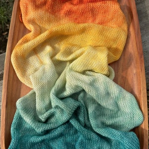 May include: A hand-knitted scarf in a gradient of orange, yellow, green, and teal colors. The scarf is folded and displayed on a wooden surface.