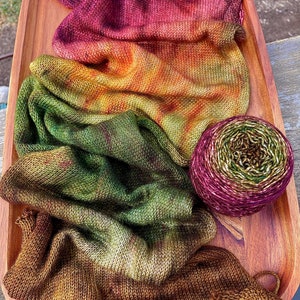 May include: A skein of hand-dyed yarn in shades of burgundy, orange, green, and brown. The yarn is arranged in a gradient pattern, with the burgundy at the top and the brown at the bottom. The yarn is displayed on a wooden tray.