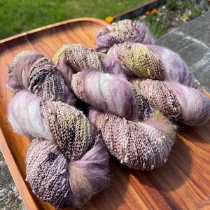 Puede incluir: Una colección de hilo teñido a mano en tonos lavanda, crema y beige. El hilo está dispuesto en una bandeja de madera, mostrando su apariencia texturizada y esponjosa. Ideal para proyectos de tejido y ganchillo.