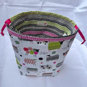 May include: A white fabric storage bag with a sheep pattern in various colors. The bag has a pink drawstring and a green and black patterned interior. The top edge is trimmed with pink zigzag fabric.