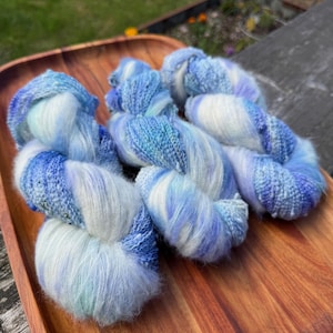 Puede incluir: Tres madejas de hilo teñido a mano en tonos azul, blanco y lavanda. El hilo está dispuesto en una bandeja de madera, mostrando su suave textura y variaciones de color. Ideal para proyectos de tejido o ganchillo.