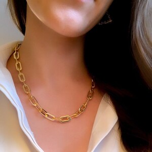 May include: A gold chain necklace with large, rectangular links. The necklace is worn by a person with a white shirt.