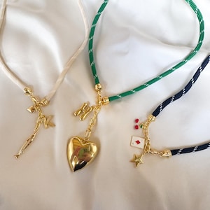May include: Three necklaces with gold-colored charms. One has a cream-colored cord with a fish and letter "K" charm. Another has a green cord with a gold heart and letter "H" charm. The third has a navy cord with a star, card, and cherry charm.