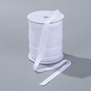 May include: A spool of white elastic ribbon. The ribbon is neatly wound around a cardboard core, with a length of the ribbon extending outwards. The ribbon is a bright white color and appears to be made of a soft, flexible material.