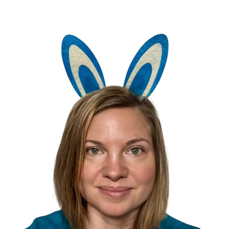 Puede incluir: Una persona con orejas de conejo azules y blancas. Las orejas son de un azul vibrante con un interior blanco y est&aacute;n unidas a una diadema. La persona tiene el pelo rubio y est&aacute; sonriendo.