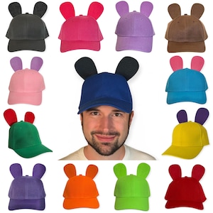 Peut inclure: Une collection de douze casquettes de baseball de différentes couleurs unies, chacune dotée de deux oreilles arrondies dressées sur la calotte. Les couleurs incluent le noir, rose, violet, marron, rose clair, bleu, vert, jaune, violet, orange, vert lime et rouge.