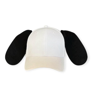 May include: A white baseball cap featuring two large, black, rounded fabric ears attached to the sides. The cap has a structured crown and a curved brim, with the black ears extending outward to resemble the appearance of a dog's floppy ears.