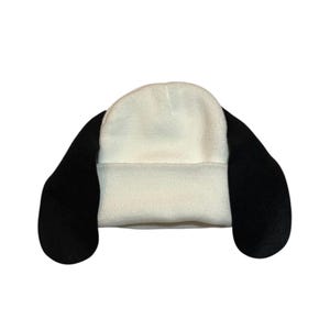 May include: A white beanie hat with black, floppy ears. The hat is made from a soft, plush material. The ears extend from the sides, giving it a playful, cartoon-like appearance.