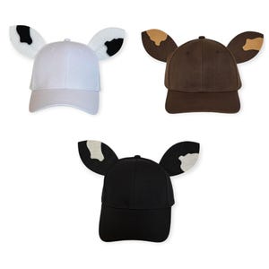 May include: Three baseball caps with animal-like ears attached to the sides. One cap is white with black-spotted ears, one is dark brown with tan-spotted ears, and one is black with white-spotted ears. Each cap features a curved brim and crown.