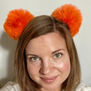 May include: A person wearing a headband with two large, fluffy, orange pom-poms. The pom-poms are round and stand out against the person's brown hair and fair skin. The background is a plain, light-colored wall.