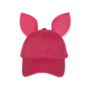 May include: A bright pink baseball cap featuring two large, upright, rounded ears attached to the top. The cap is constructed from a solid-colored fabric with a structured crown, a curved brim, and visible stitching details throughout the design.
