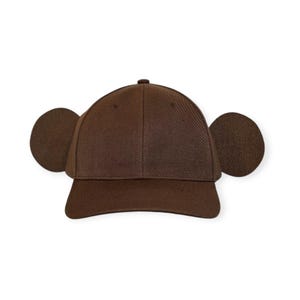 Puede incluir: Una gorra de béisbol de color marrón oscuro sólido con una corona estructurada y una visera curva. La gorra está decorada con dos orejas de tela grandes, circulares y de color marrón oscuro a cada lado, que se extienden hacia afuera.