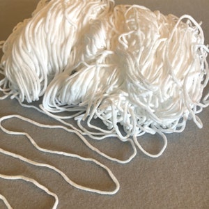 May include: A large pile of white elastic cord, commonly used for face masks, with a single cord laid out in a loop on a gray surface.