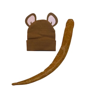May include: A brown knit beanie featuring two attached animal ears with light pink inner accents, paired with a long, curved, textured brown tail accessory. The beanie has a cuffed brim and a solid brown color throughout the main fabric.