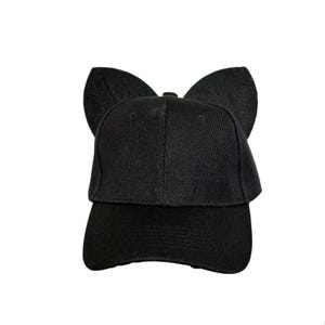 May include: A solid black baseball cap featuring two prominent, upright, rounded felt-like ears attached to the top of the crown. The cap has a structured front panel, a curved brim, and a consistent matte black fabric finish throughout.