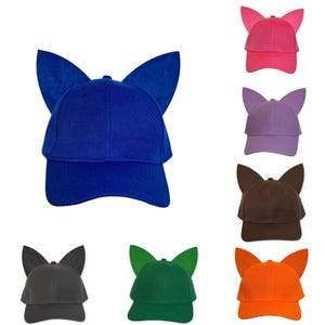 May include: A collection of baseball caps featuring prominent, upright cat-like ears attached to the crown. The caps are available in various solid colors including royal blue, hot pink, lavender, dark brown, charcoal gray, forest green, and orange.