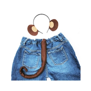 Può includere: Un accessorio per costume di coda di scimmia marrone con una fascia per capelli con orecchie marroni con centri beige. La coda è attaccata a un paio di pantaloncini di jeans blu.
