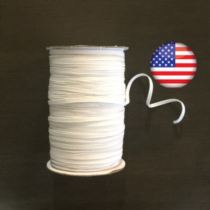 May include: A spool of white elastic cord, wound on a cardboard tube, with a small piece of the cord extending out. The spool is sitting on a dark brown surface. A small American flag sticker is in the background.