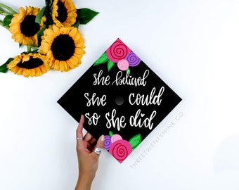 Può includere: Un cappello di laurea nero decorato con fiori rosa, viola e verdi. Il cappello ha una scritta bianca che recita "She believed she could so she did" e il sito web "THREETWENTYNINE.CO".
