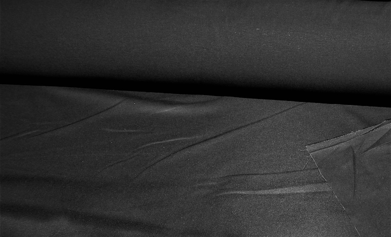 May include: A roll of black fabric, with visible folds and a slightly textured surface. A cut piece of the same fabric lies to the right, showing the raw edge. The fabric appears to be a solid color, suitable for various sewing projects.