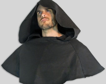 May include: A black hooded cloak with a long, flowing cape. The hood is pulled up over the head, and the cloak is worn over a plain white shirt.