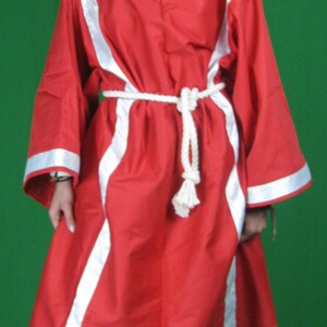 May include: A woman wearing a red robe with white trim and a white rope belt. The robe has a long, flowing design and is cinched at the waist. The woman is standing on a green background.