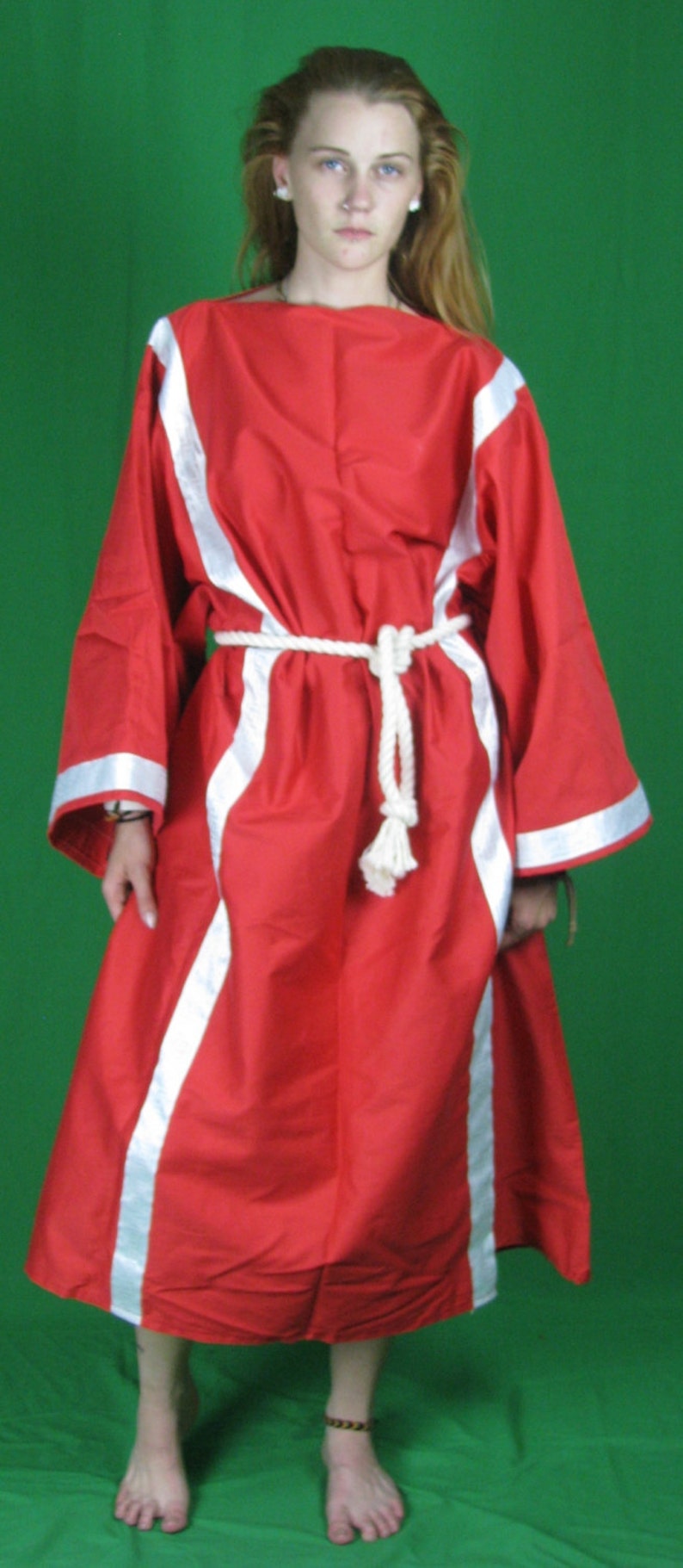 May include: A woman wearing a red robe with white trim and a white rope belt. The robe has a long, flowing design and is cinched at the waist. The woman is standing on a green background.