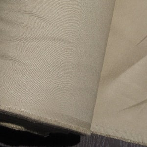 May include: A roll of beige fabric, tightly wound, with a visible textured weave. The fabric is unhemmed at the edge, revealing a raw, frayed texture. The color is a neutral, light tan.