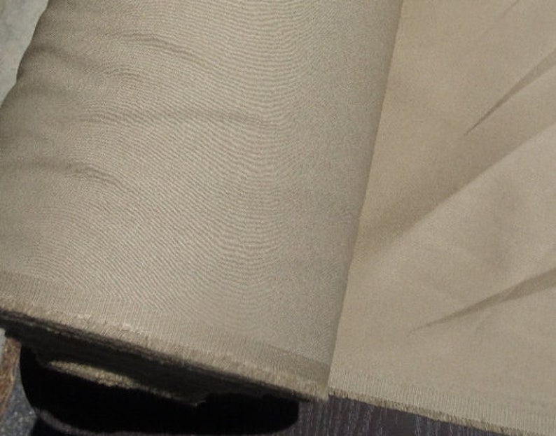 May include: A roll of beige fabric, tightly wound, with a visible textured weave. The fabric is unhemmed at the edge, revealing a raw, frayed texture. The color is a neutral, light tan.