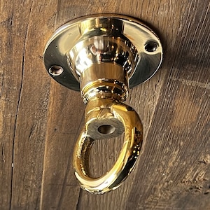 May include: A gold-toned metal ceiling hook with a round base and a ring at the bottom. The hook is mounted on a wooden surface.