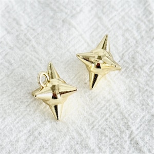 May include: Two gold-colored star-shaped charms. Each charm has four pointed tips and a loop for attaching to jewelry. The charms are displayed on a white, textured surface.