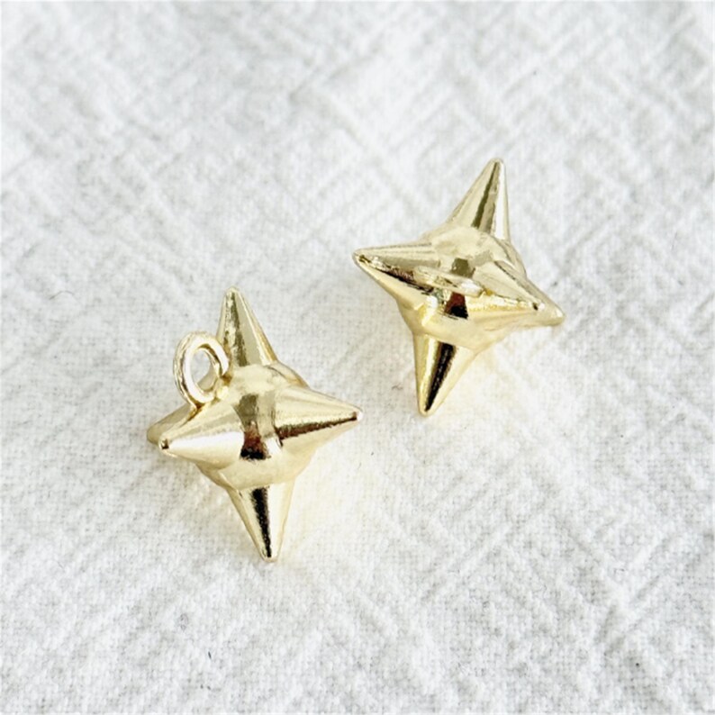 May include: Two gold-colored star-shaped charms. Each charm has four pointed tips and a loop for attaching to jewelry. The charms are displayed on a white, textured surface.