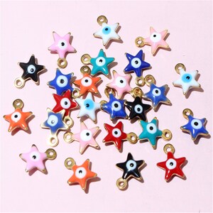 May include: A collection of star-shaped charms in various colors, including pink, blue, orange, and black. Each charm features a central eye design and a gold-colored loop for attachment. The charms are scattered on a pink background.