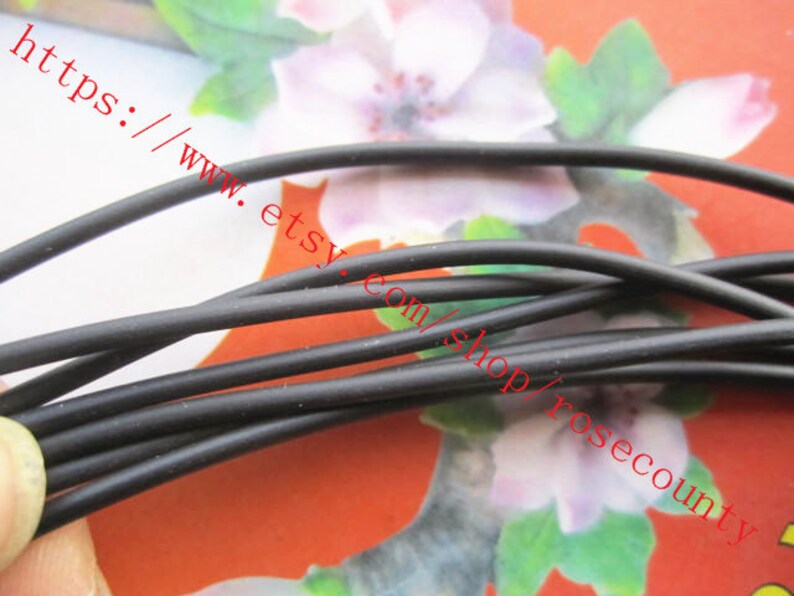 Puede incluir: Un manojo de cord&oacute;n negro fino y flexible con un acabado mate suave. Las hebras est&aacute;n dispuestas en un grupo suelto y superpuesto, mostrando una forma cil&iacute;ndrica consistente a lo largo de todo el material sobre un fondo floral.