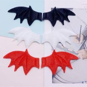 May include: A collection of six felt bat wing appliques in three colors: black, white, and red. Each pair of wings is designed with detailed veining and a curved shape, perfect for crafting or costume embellishments.