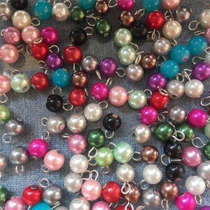 May include: A collection of pearl beads in various colours, including white, pink, green, blue, and black. The beads are strung on silver wire loops, creating a decorative and versatile craft supply.